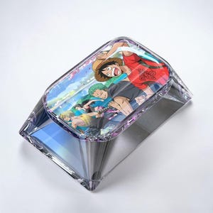 May include: A clear, rectangular acrylic box with a hinged lid. The lid features a colorful anime illustration of several characters. The box has a prismatic effect, reflecting light. The design is likely inspired by the anime series "One Piece."