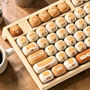 Puede incluir: Un teclado único con teclas personalizadas. Las teclas están diseñadas para parecerse a pan y bollos con caras lindas. El teclado tiene un marco beige e incluye teclas de función y teclas especiales como "ESC", "TAB" y "SHIFT".