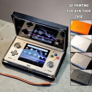 Puede incluir: Consola de juegos portátil blanca y plateada, con una pantalla que muestra iconos de aplicaciones. Está en una funda negra, con una correa marrón. El texto "3D PRINTING FOR AYN THOR CASE" es visible. Se muestran fundas adicionales.