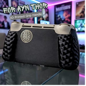 May include: A black and beige 3D printed grip handle for the AYN THOR handheld gaming device. The handle features textured grips, a circular vent, and the text "FOR AYN THOR" and "3D Printed Grip Handle".