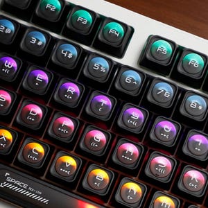 Könnte beinhalten: Nahaufnahme einer mechanischen Tastatur mit schwarzen Tasten und einem weißen Rahmen. Die Tasten haben farbige, hintergrundbeleuchtete Buchstaben und Symbole. Die Funktionstasten F2 bis F6 sind sichtbar. Die Tastatur hat die Aufschrift "SPACE RX-1(o)".