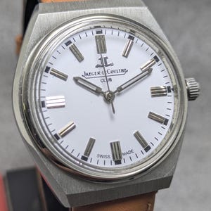 May include: A stainless steel Jaeger-LeCoultre Club wristwatch with a white dial, silver-toned baton hour markers, and silver hands. The watch features a brushed metal case, a crown on the right side, and the text Jaeger-LeCoultre Club and Swiss Made.
