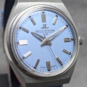 Könnte beinhalten: Eine Jaeger-LeCoultre Club Armbanduhr mit hellblauem Zifferblatt, silberfarbenen Stundenmarkierungen und silbernen Zeigern. Die Uhr hat ein Gehäuse aus gebürstetem Edelstahl und ein hellblaues, strukturiertes Lederarmband. Das Zifferblatt zeigt den Markennamen und Swiss Made.