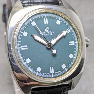 Puede incluir: Un reloj Breitling Geneve vintage con caja plateada y esfera verde oscuro. El reloj presenta agujas e indicadores plateados y una correa de cuero negro. La esfera indica "Breitling Geneve" y "Swiss Made".