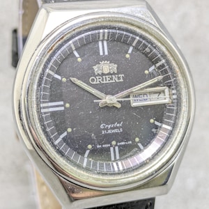 May include: A silver-toned Orient watch with a black face. The watch face displays the Orient logo, the day and date, and the words "Crystal" and "21 Jewels." The watch has a black leather band.