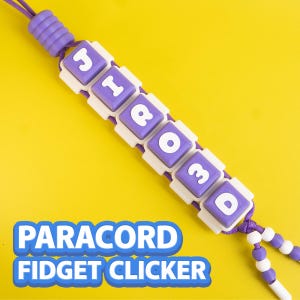 Pode incluir: Um cordão de paracord roxo com cinco contas quadradas estilo tecla de teclado, com base branca e topo roxo, exibindo as letras brancas J, I, R, 3 e D. O cordão inclui uma conta roxa estriada no topo e contas menores roxas e brancas.