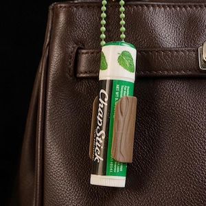 Op de afbeelding: Een ChapStick lippenbalsemstick met een groen-wit etiket met illustraties van muntblaadjes, vastgehouden door een bruine, getextureerde hoes bevestigd aan een lichtgroene kogelketting, hangend tegen een donkerbruine handtas van generfd leer.