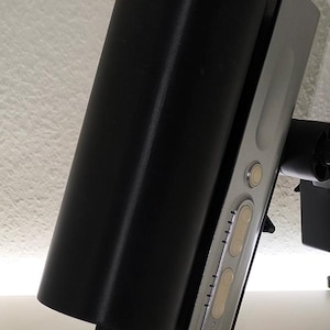 Könnte beinhalten: Ein schwarzes zylindrisches Objekt ist an einer silberfarbenen rechteckigen Basis mit mehreren Knöpfen befestigt. Das Objekt ist leicht abgewinkelt, wobei sich der zylindrische Teil über der Basis befindet. Der Hintergrund ist eine schlichte weiße Oberfläche.