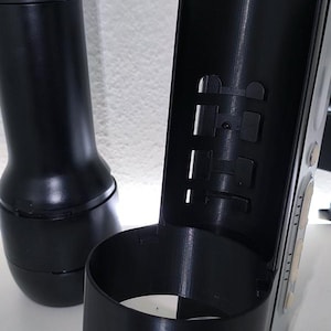 Könnte beinhalten: Zwei schwarze Kunststoffobjekte. Eines ist zylindrisch, das andere ist eine höhere, offene Komponente mit rechteckigen Aussparungen. Die Objekte befinden sich auf einer weißen Oberfläche.