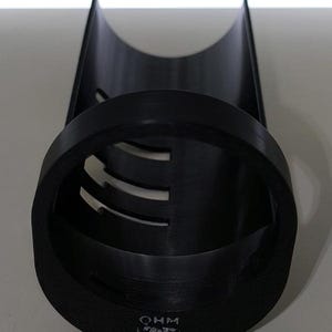 May include: A black cylindrical object with a rounded top and a circular base. The base has the letters "OHM" printed on it. The cylinder has several horizontal cutouts. The object is made of a matte black material.