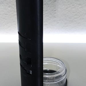 May include: A black, U-shaped holder with cut-out designs, paired with a clear glass jar. The jar has a silver lid with text. The holder is made of a matte material and stands upright on a white surface.