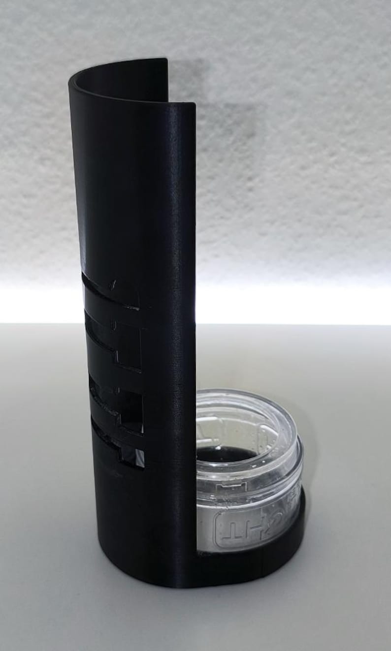 May include: A black, U-shaped holder with cut-out designs, paired with a clear glass jar. The jar has a silver lid with text. The holder is made of a matte material and stands upright on a white surface.