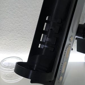 May include: A black and silver electronic device with a curved black holder attached. The holder has rectangular cutouts and a small shelf. A clear glass jar with white text is in the background.