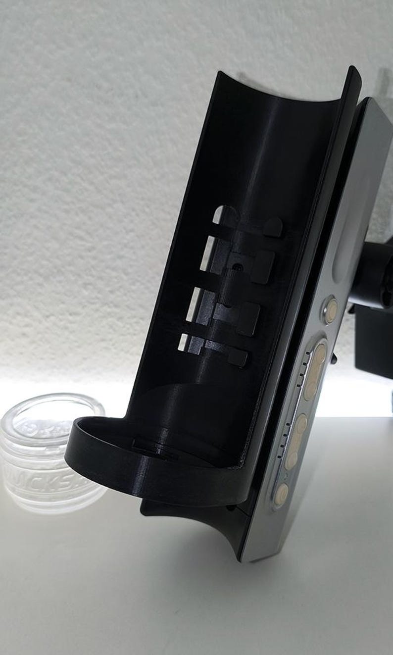 May include: A black and silver electronic device with a curved black holder attached. The holder has rectangular cutouts and a small shelf. A clear glass jar with white text is in the background.