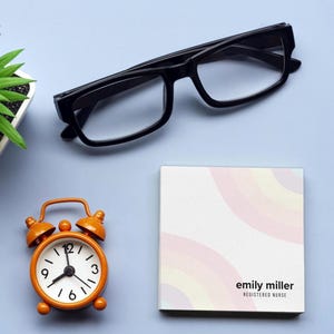 May include: Black-framed eyeglasses, an orange alarm clock, and a notepad with a pastel rainbow design and the text "emily miller REGISTERED NURSE" are arranged on a light blue surface. A small potted plant is visible.
