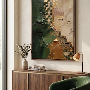 May include: Abstract framed artwork with textured brushstrokes in shades of green, gold, and beige. The painting features geometric patterns and a rich, layered appearance, creating a modern and artistic aesthetic. The artwork is displayed in a room with a wooden cabinet and a green armchair.