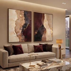 May include: Two large square framed wall art pieces featuring abstract layers of cream, beige, deep burgundy, and black paint with gold leaf accents. The paintings hang above a beige sofa with dark red and brown throw pillows and a marble coffee table.