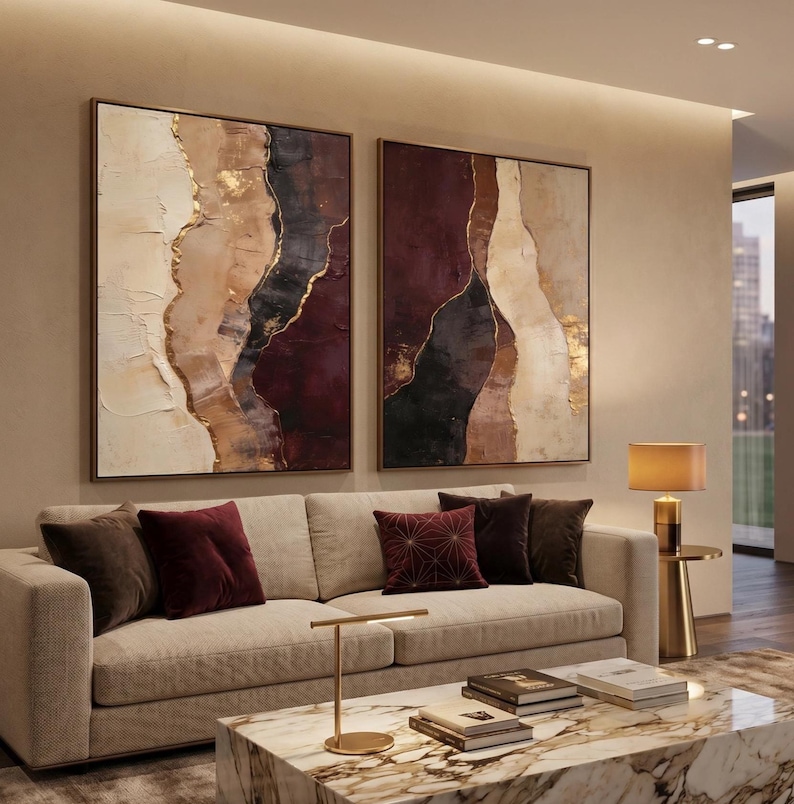 May include: Two large square framed wall art pieces featuring abstract layers of cream, beige, deep burgundy, and black paint with gold leaf accents. The paintings hang above a beige sofa with dark red and brown throw pillows and a marble coffee table.