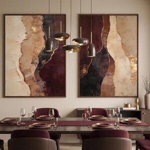 May include: Two large rectangular abstract paintings featuring layered textures in shades of cream, beige, deep burgundy, and black with gold leaf accents, displayed above a dining table set with burgundy chairs and a dark wood table.