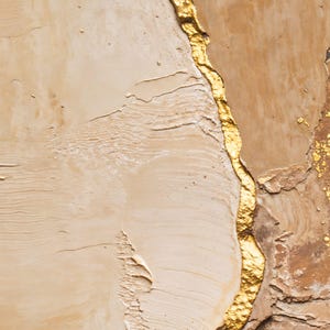May include: An abstract painting featuring thick, textured layers of beige and tan paint. A prominent, jagged vertical line of metallic gold leaf runs through the center, with additional gold leaf accents scattered on the right side of the canvas.