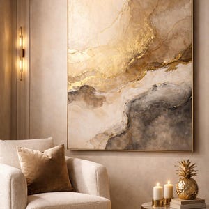 Peut inclure: Tableau abstrait aux tons or et neutres, encadré d'or. Un fauteuil crème avec un coussin en velours marron est en dessous. Une table d'appoint dorée présente des bougies et un ananas décoratif. L'œuvre est un point focal.