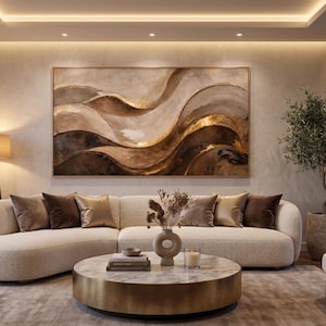May include: A contemporary living room with a large abstract artwork in brown and gold hues above a curved beige sofa. A round coffee table with a marble top and gold base is in front of the sofa, with decorative items.