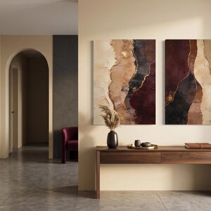 May include: Two rectangular wall art canvases featuring abstract vertical layers of deep burgundy, charcoal black, cream, and beige with metallic gold accents. A dark vase with dried pampas grass, a gold tray with small bowls, and a book sit on a wooden console.