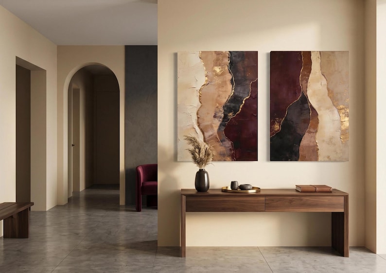 May include: Two rectangular wall art canvases featuring abstract vertical layers of deep burgundy, charcoal black, cream, and beige with metallic gold accents. A dark vase with dried pampas grass, a gold tray with small bowls, and a book sit on a wooden console.