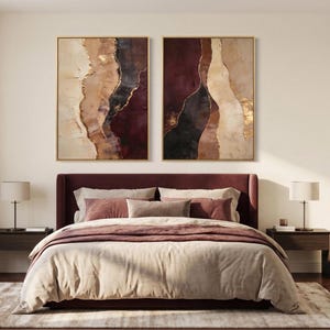 May include: Two rectangular wall art pieces featuring abstract vertical layers of cream, tan, deep burgundy, and black paint with metallic gold accents, displayed in thin gold frames above a dark burgundy upholstered bed with neutral bedding.