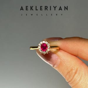 Könnte beinhalten: Ein goldener Ring mit einem zentralen ovalen Rubin, umgeben von einem Kranz aus kleinen Diamanten. Der Ring wird vor einem neutralen Hintergrund gehalten. Der Markenname "AEKLERIYAN JEWELLERY" ist oben angezeigt.