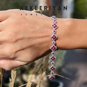 May include: A diamond bracelet with ruby accents. The bracelet features a series of four-leaf clover-shaped designs, each with a central ruby surrounded by smaller diamonds. The bracelet is worn on a wrist, with the brand name "AEKLERIYAN JEWELRY" visible.