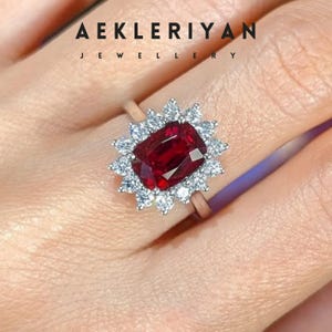 May include: A ring featuring a large, rectangular, deep red gemstone surrounded by a halo of smaller, clear stones. The ring's band is a light gold color. The text "AEKLERIYAN JEWELLERY" is at the top of the image.
