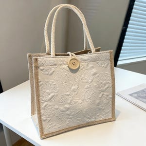 May include: Cream-colored tote bag with a floral embossed design. The bag features a button closure and rope handles. The edges are trimmed with a natural-colored fabric. The bag is sitting on a white surface.