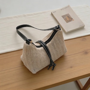 May include: A beige woven handbag with a black leather strap and accents. The bag has a rectangular shape and a zipper closure. A magazine with a pear illustration is next to the bag on a wooden surface.