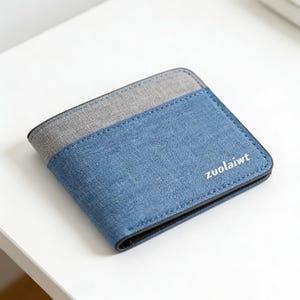 Puede incluir: Una cartera azul y gris de dos pliegues con el texto "zuolaiwt" en blanco. La cartera tiene un aspecto texturizado y un borde negro. La cartera está sobre una superficie blanca.