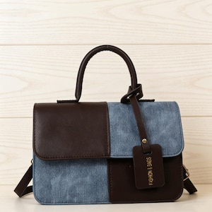 Puede incluir: Un bolso de mano de dos tonos con un asa marrón y una correa para el hombro desmontable. El bolso presenta un diseño de bloques de color con paneles marrones y azules. Una pequeña etiqueta marrón con el texto "FASHION & BAGS" cuelga del asa.