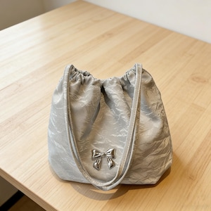Puede incluir: Un bolso con cordón gris claro con un patrón texturizado y un lazo plateado. El bolso tiene una parte superior fruncida y dos correas. El bolso está sobre una superficie de madera marrón claro.
