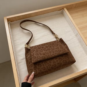 Könnte beinhalten: Braune Schultertasche mit Leopardenmuster. Die Tasche hat einen Klappenverschluss und einen langen Riemen mit goldfarbenen Kettenakzenten. Die Tasche liegt auf einer hellen Oberfläche.