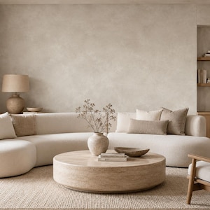 May include: A curved cream-colored sofa with textured fabric, paired with a round beige stone coffee table. The table holds a ceramic vase with dried branches and a shallow bowl. A wooden armchair with cream cushions sits to the right.