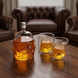 May include: A clear glass decanter and two matching glasses, all filled with amber liquid. The decanter and glasses are shaped like a Stormtrooper helmet. The set is displayed on a dark wooden table, suggesting a bar or home entertainment setting.