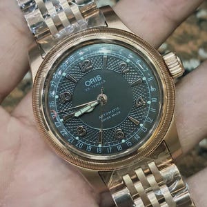 Puede incluir: Un reloj Oris dorado con esfera verde oscuro y correa dorada. La esfera del reloj muestra la marca "ORIS" y el texto "25 JEWELS". El reloj tiene una pantalla de fecha y es un reloj automático de fabricación suiza.
