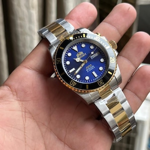 May include: A wristwatch with a blue face, gold accents, and a silver and gold band. The watch has a black bezel with white markings and a date display. The watch is held in a hand.