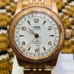 Può includere: Un orologio Oris color oro rosa con quadrante bianco e visualizzazione della data. L'orologio presenta un quadrante strutturato con numeri e lancette nere. Il cinturino dell'orologio è anch'esso color oro rosa.