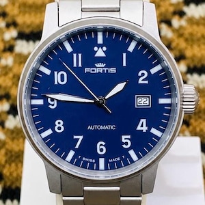 May include: A stainless steel Fortis automatic watch with a deep blue face. The watch features white numerals, hands, and a date display at the 3 o'clock position. The watch band is made of stainless steel.