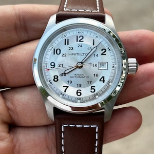 May include: A Hamilton Khaki Automatic wristwatch with a silver dial, black Arabic numerals, and a 24-hour inner track. The watch features a date window at the three o'clock position, luminous hands, and a brown leather strap with white stitching.