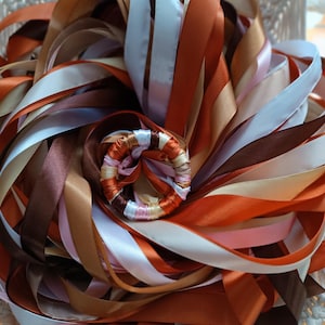 Peut inclure: Gros plan sur un arrangement décoratif de rubans. Les rubans sont dans des tons de marron, blanc, orange et rose. Les rubans sont disposés en cercle, avec un petit arrangement circulaire au centre. Le fond est une texture tissée de couleur crème.