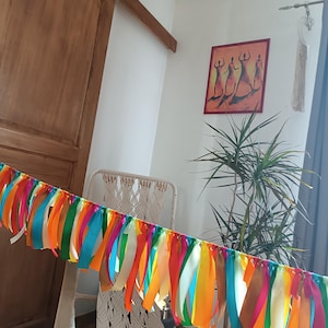 May include: A decorative garland featuring a series of hanging ribbons in vibrant shades of orange, yellow, pink, green, and turquoise. The ribbons are attached to a horizontal cord, creating a colourful fringe effect suitable for festive decor.