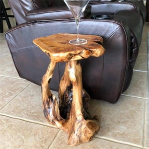 May include: A unique, handcrafted side table made from natural wood. The table features a live edge top and a base constructed from intertwined branches. A clear martini glass sits on the table's surface. The wood has a warm, natural finish.