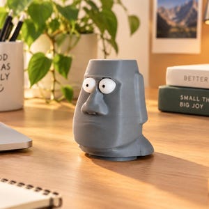 Könnte beinhalten: Ein grauer, 3D-gedruckter Stiftehalter oder Übertopf in Form eines Moai-Kopfes mit großen, weit aufgerissenen Cartoon-Augen. Die Figur hat eine flache Öffnung an der Oberseite, eine markante Nase und einen neutralen Gesichtsausdruck.