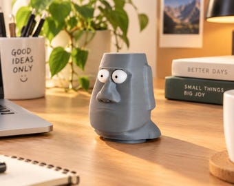 May include: A gray, 3D-printed Moai-style head planter or desk organizer featuring large, wide-eyed cartoonish eyes. The figure has a flat top opening, a prominent nose, and a neutral facial expression, standing upright on a flat surface.